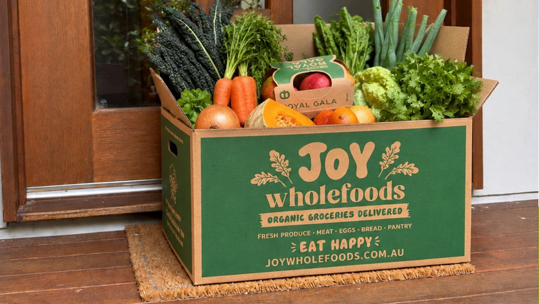 Joy Wholefoods organic fruit and vegetable delivery box filled with fresh seasonal produce, including leafy greens, carrots, apples, citrus, pumpkin and herbs, sitting on a doorstep ready for delivery.