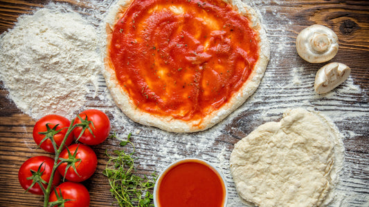 Homemade pizza dough topped with rich tomato sauce, surrounded by fresh tomatoes, mushrooms, herbs and pizza ingredients on a wooden table.