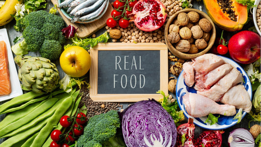 Organic fruit and vegetables, pasture-raised chicken, wild-caught salmon, nuts, seeds surrounding a small blackboard that says 'Real Food'