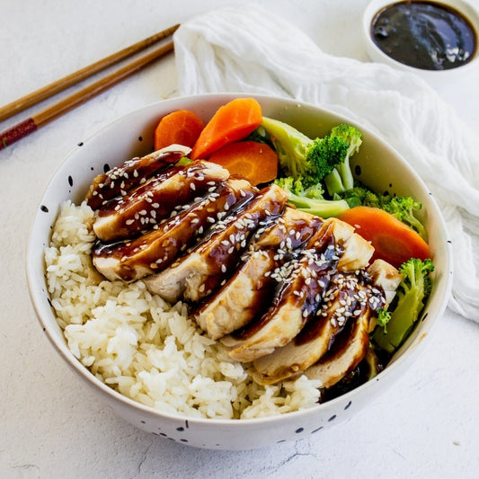 Bowl of rice with teriyaki chicken, carrots, and broccoli on a light surface.