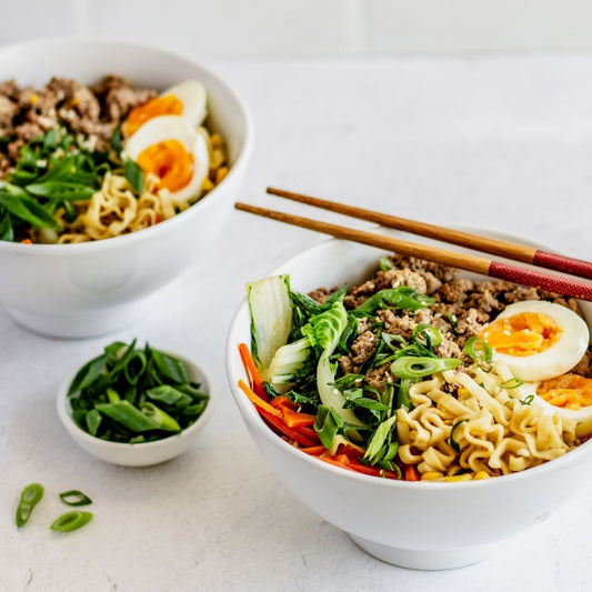 Two bowls of ramen with vegetables and eggs on a white surface