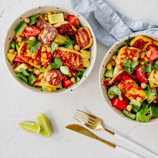 Two bowls of colorful salad with halloumi cheese on a light surface with cutlery.