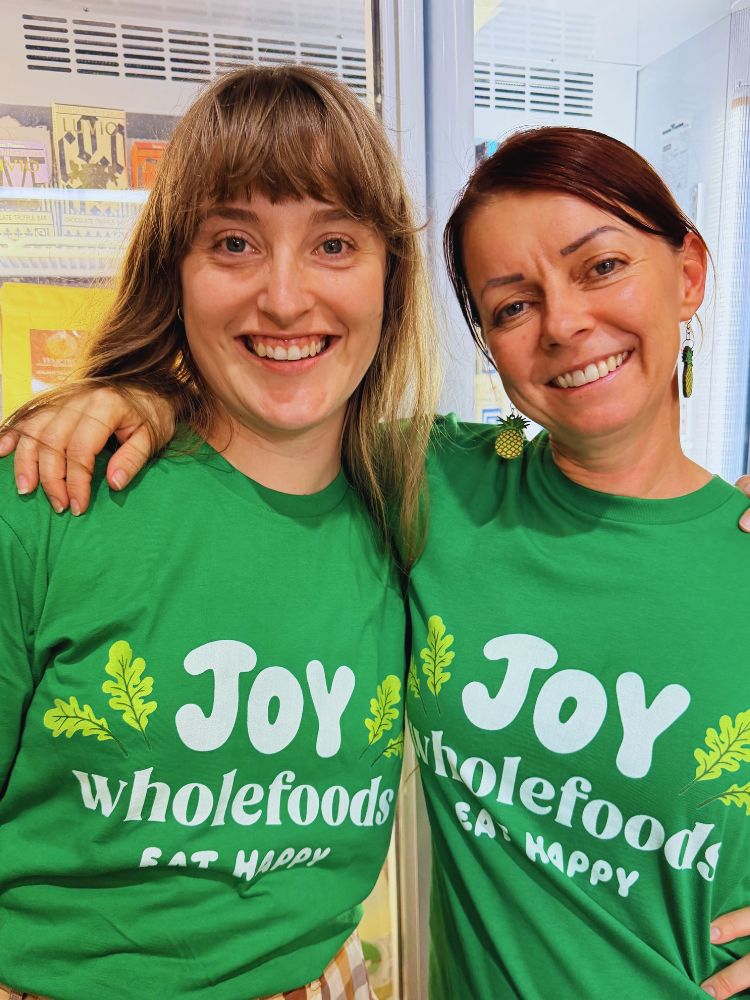 Two women wearing green 'Joy Wholefoods' t-shirts in a store setting.