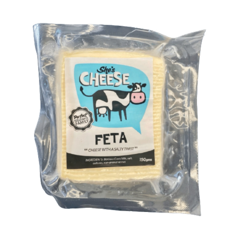 A packaged block of feta cheese in a clear plastic bag with a printed front label reading 'She's Cheese' and 'FETA'. The cheese itself is pale yellow, and the packaging is transparent, showing the product clearly against a plain white background.