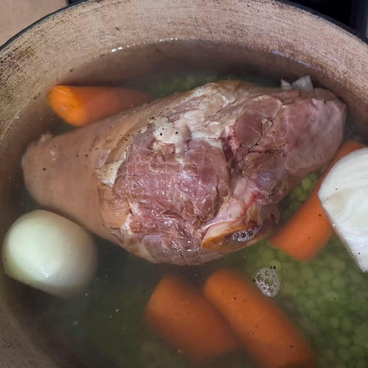 A close-up of a large raw pork shoulder simmering in a pot with carrots, a white onion, and green peas in clear broth.