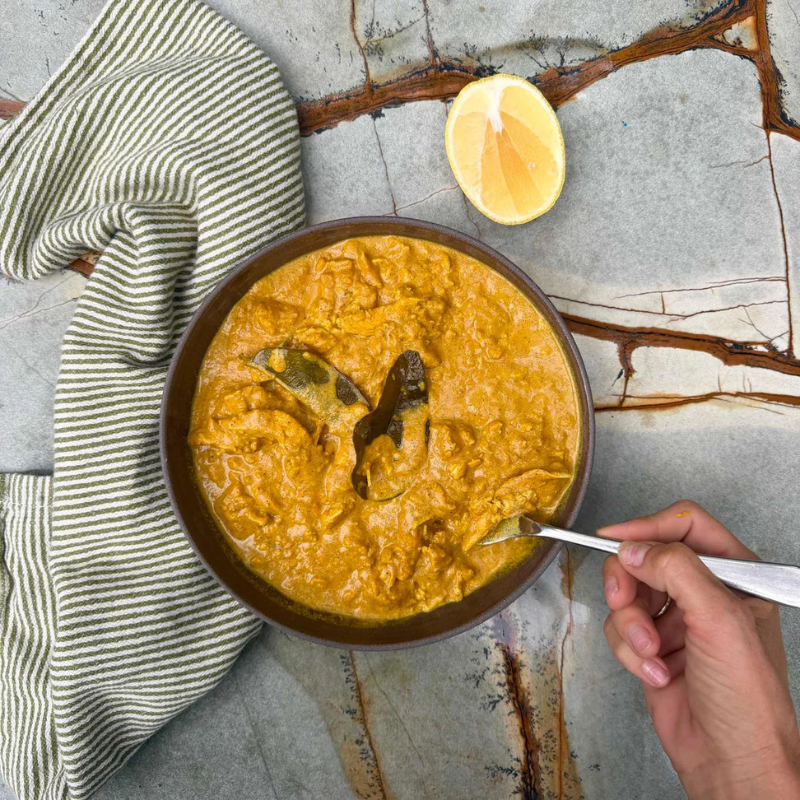 Pumpkin soup in a bowl with a hand holding a spoon, lemon wedge, and striped towel on a textured surface.