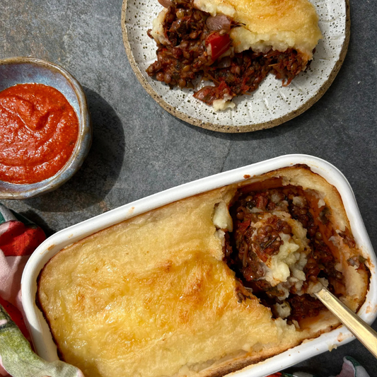 A baked shepherd's pie casserole in a white rectangular baking dish, with a spoon lifting a portion to show the meat and mashed potato filling. A smaller plated slice sits in the background, and a small bowl of red sauce is on the left, all on a dark gray surface.