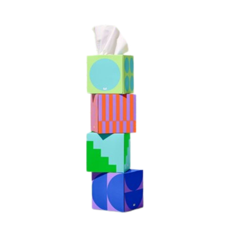 A stack of four colorful geometric tissue boxes on a plain white background. The top box is light green with a large light blue circle, with a white tissue protruding.
