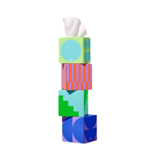 A stack of four colorful geometric tissue boxes on a plain white background. The top box is light green with a large light blue circle, with a white tissue protruding.